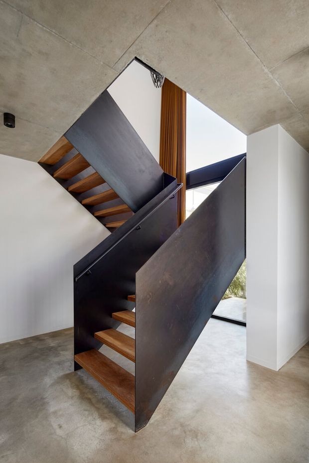 A sculptural interior stairway with dark metal plate balustrades and stringers, warm timber treads, polished concrete flooring, white plaster walls, and an exposed concrete ceiling. Vertical timber screening and large glazing add contrast and natural light to the minimalist double-height circulation space.