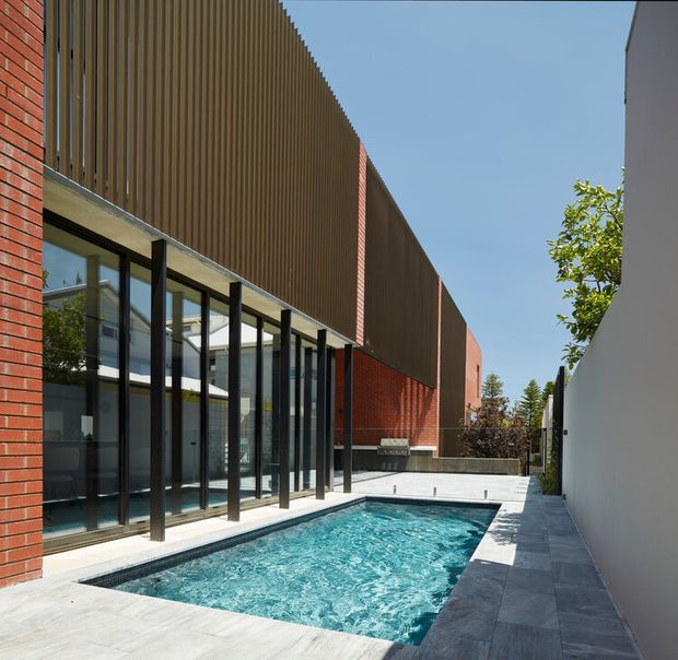 Contemporary architectural courtyard with a rectangular swimming pool beside a brick-and-metal building. Full-height black-framed glazing opens onto pale paved pool surrounds, while vertical ribbed screening forms a strong upper facade element. A narrow white boundary wall and landscaped planting define the compact outdoor space under a clear sky.