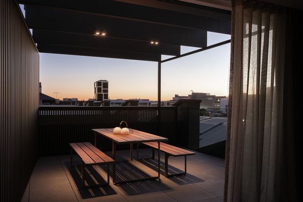 A covered rooftop or upper-level terrace with a timber picnic-style dining table and benches, enclosed by dark vertical screening and a partially open metal-framed roof. Sheer curtains frame the right side, while an urban skyline is visible beyond the balustrade at sunset. The scene has a dark, contemporary architectural character with warm low-angle light.