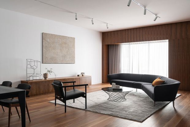 A refined mid-century-inspired living room with dark timber wall panelling and built-in cabinetry, warm wood flooring, a large black curved sofa, matching lounge chair, low wire coffee table, textured area rug, and minimalist track lighting. Sheer curtains soften the large window, while abstract artwork and sparse decor create a calm, sophisticated atmosphere.