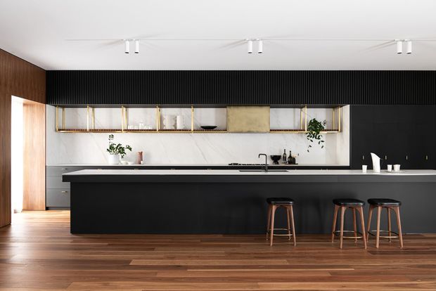 Contemporary kitchen with a large matte-black island and three timber bar stools, dark vertical fluted wall detailing, white marble-look backsplash, open shelving with brass-toned supports, integrated cabinetry, timber flooring, and minimalist track lighting.