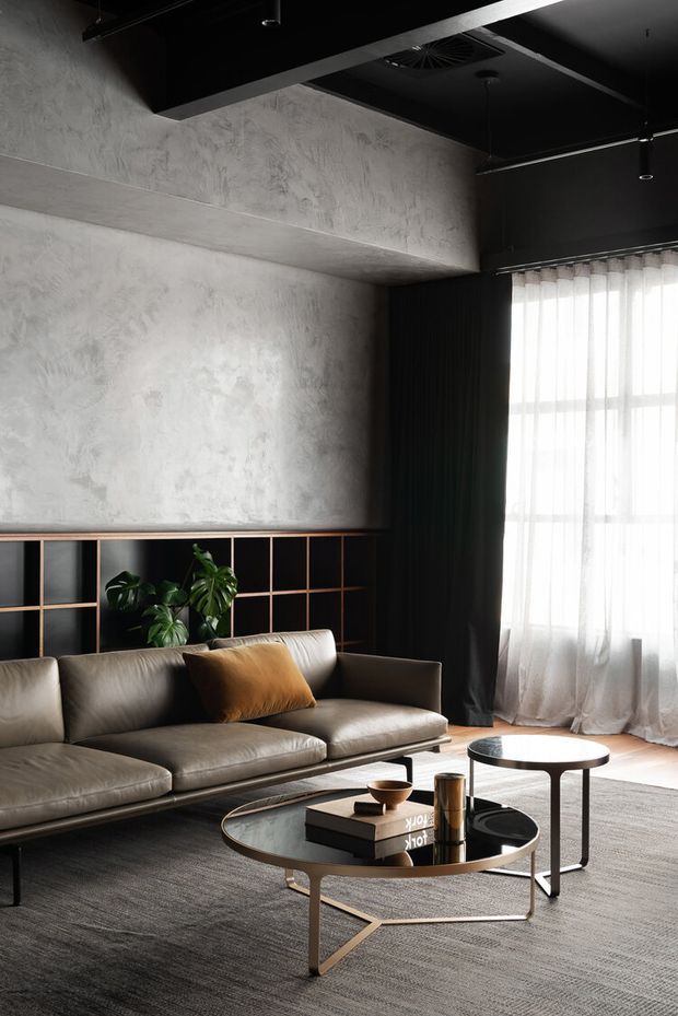 A sophisticated contemporary living room with a dark ceiling, textured grey concrete-look walls, and a large sheer-curtained window. A low-profile taupe leather sectional sofa sits against built-in black shelving with warm timber trim and indoor greenery. Two round coffee tables, a textured grey rug, and restrained styling create a moody, refined atmosphere.