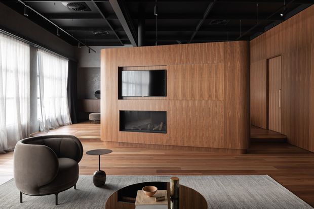 A sophisticated open-plan living room with a warm timber feature wall incorporating a television and linear fireplace. The room combines dark exposed ceiling services and beams with floor-to-ceiling sheer curtains, timber flooring, a grey area rug, a curved dark upholstered lounge chair, and a low round coffee table. The overall composition is refined, warm, and contemporary with strong natural wood grain and high contrast between the dark ceiling and lighter finishes.