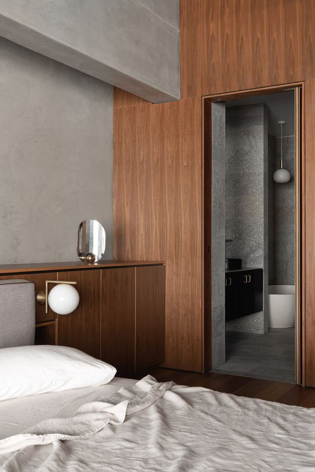 A sophisticated bedroom with a partially visible bed in the foreground, dark walnut-toned timber wall panelling and built-in cabinetry, a concrete-textured wall and ceiling beam, and sculptural globe lighting. An open doorway leads directly to a compact ensuite bathroom finished in dark grey stone-like surfaces, with black cabinetry and a white toilet visible.