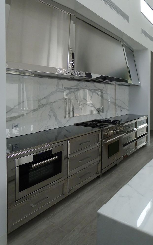 A sleek contemporary kitchen with a full wall of stainless-steel cabinetry, integrated appliances, a large gas range, and a dramatic white-and-grey veined marble backsplash. A white island is partially visible in the foreground. The space has a refined monochromatic palette and glossy reflective finishes.