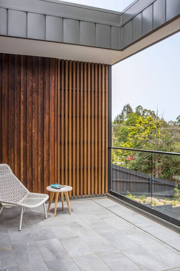 A modern covered outdoor terrace or balcony with large grey stone-look floor tiles, a dark timber-clad wall, and a vertical timber slatted screen. The space includes a woven lounge chair and small round timber side table, with dark metal balustrading and views toward a landscaped suburban garden. The roof edge and upper wall panels are finished in grey metal, creating a refined contemporary architectural composition.