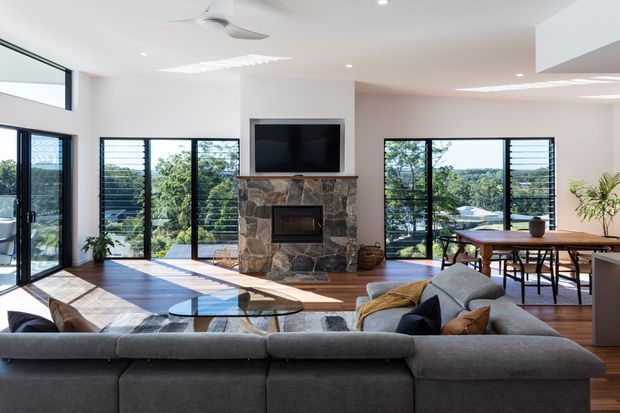 Bright open-plan living and dining area with a large grey sectional sofa in the foreground, a glass coffee table, timber flooring, extensive black-framed louvred glazing, and a feature natural-stone fireplace with built-in television. A dining table and chairs sit beside the windows, with leafy views beyond.