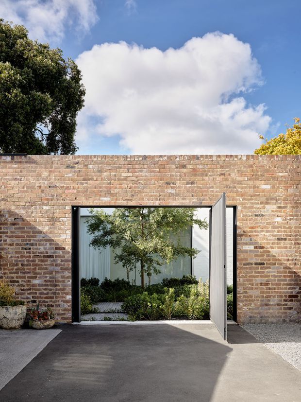A contemporary courtyard or garden entrance viewed from outside, set within a weathered red-brick boundary wall. A large black-framed glazed opening with a pivoting metal door leads into a planted courtyard, while paved and gravel surfaces occupy the foreground. Mature trees, blue sky and clouds are visible above the wall.