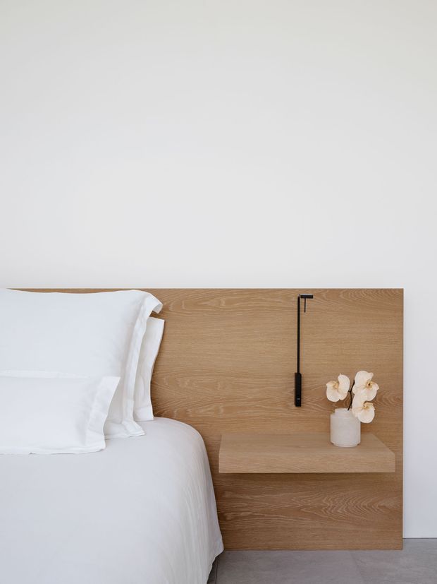 Minimalist bedroom detail showing a white bed against a full-height light timber headboard with an integrated floating bedside shelf and slender black wall-mounted reading light. A small white vase with pale flowers adds a subtle decorative touch. The palette is soft white, light wood, and pale grey.