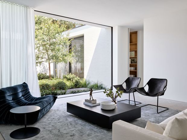 A bright, minimalist living room with floor-to-ceiling glazing opening onto a landscaped courtyard. The room features a dark sculptural lounge chair, two black leather sling chairs, a white sofa, a low black rectangular coffee table, soft grey carpet, sheer curtains, and a recessed timber display bookshelf. White walls and abundant natural light create a refined contemporary setting.