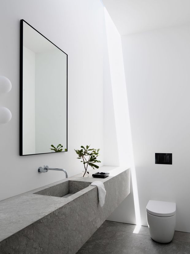 A minimalist contemporary bathroom featuring a monolithic grey stone vanity with an integrated rectangular basin, wall-mounted chrome tapware, a frameless black-edged mirror, concealed black flush plate, and a compact white toilet. Bright white walls and a strong diagonal shaft of natural light create a stark, sculptural atmosphere, with a small plant adding an organic accent.