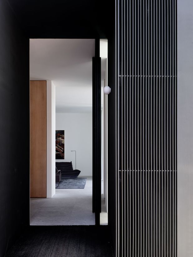 A dark, minimalist entry passage framed by black surfaces, with a tall black-framed doorway leading into a bright living room. The entrance features a striking full-height vertical metal slat screen, while the interior beyond has pale walls, a warm timber built-in cupboard, grey flooring, a dark lounge chair and a muted rug.