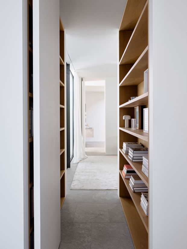 A narrow, bright interior passage lined with built-in timber shelving and books. White walls and ceiling contrast with warm wood joinery, while large glazing with sheer curtains brings in natural light. The hallway leads toward a light-filled room with pale tiled or stone flooring, creating a clean, minimalist and refined atmosphere.