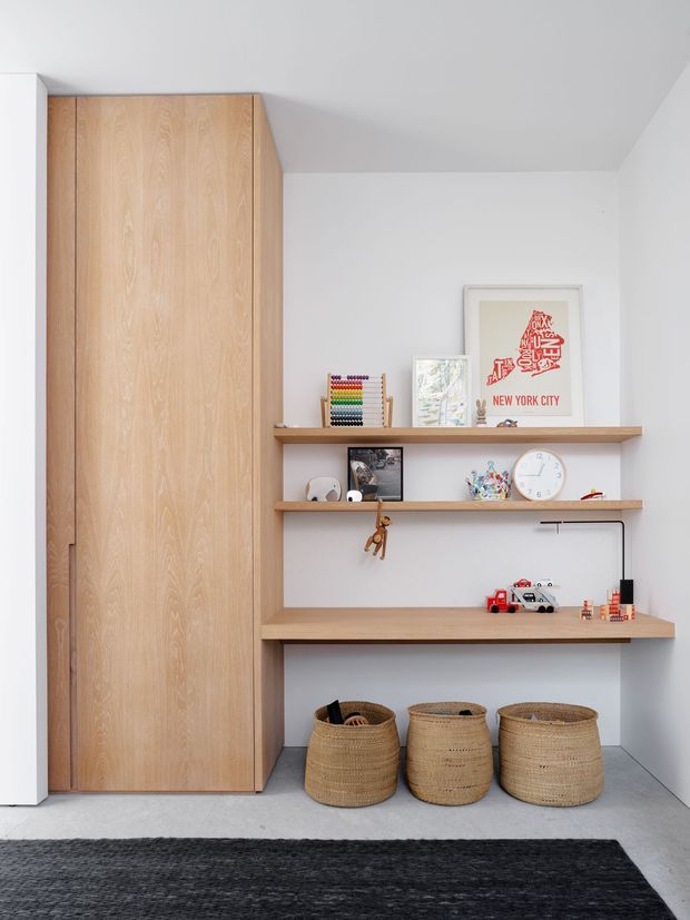 A bright, minimalist built-in children’s storage and study nook. Full-height light timber cabinetry sits beside open timber shelves and a desk-like work surface, styled with books, toys, artwork, baskets and a small lamp. White walls and a pale grey floor create a clean, contemporary setting, with a dark rug in the foreground.