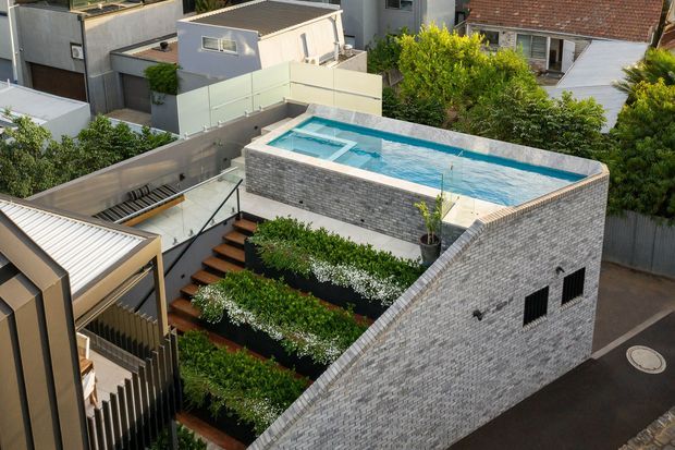 Elevated exterior view of a compact urban property featuring a rooftop swimming pool enclosed by pale grey brick walls. Timber steps and layered planted garden beds lead up beside the pool, with neighbouring homes and rooftop structures visible around the site. The design has a contemporary, space-efficient character with strong geometric forms and integrated landscaping.