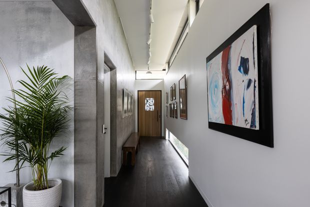 A long, narrow contemporary hallway with dark timber flooring, smooth white walls, a grey concrete-textured structural column and wall, recessed linear ceiling lighting, and a timber front door at the far end. Framed abstract artwork lines the right wall, while a tall potted palm sits near the entrance.