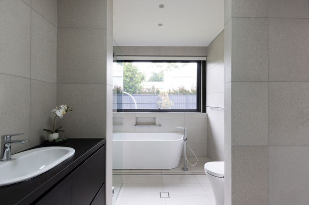 A bright contemporary bathroom viewed through a partial opening, featuring large pale grey wall tiles, matching light floor tiles, a dark floating vanity with white basin, a freestanding white bathtub beneath a wide black-framed window, and a white toilet. Minimal recessed ceiling lights and clean-lined fixtures create a calm, refined feel.