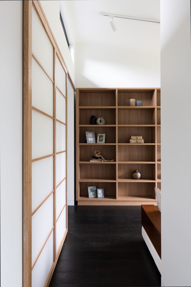 A bright, narrow interior passage or room featuring a floor-to-ceiling timber display shelving unit with open cubbies and warm wood grain. A large sliding or partition screen with a light timber grid and translucent white panels occupies the left side. The space has dark timber flooring, white walls and ceiling, and minimalist track lighting. Small decorative objects and books are arranged on the shelving, with part of a bench or upholstered furniture visible at right.