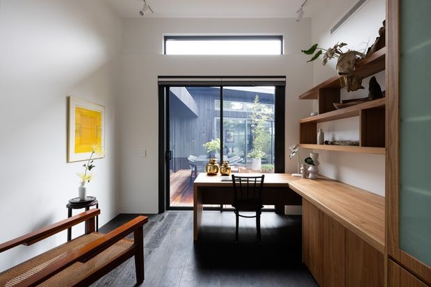 A bright, compact home office or study with a built-in timber desk and shelving along the right wall, a central desk facing a full-height glazed door, and a black chair. Dark timber flooring, white walls, track lighting, framed artwork, and indoor plants create a warm contemporary workspace. A small outdoor courtyard is visible beyond the glazing.