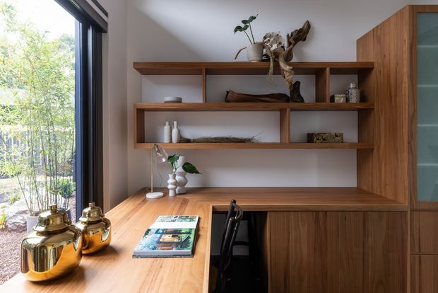 A warm, compact built-in home office nook beside a large glazed opening. The space features a timber desk integrated with extensive wall shelving and cabinetry, with open display shelves, a dark-framed window or door, books, ceramics, plants and natural sculptural decor. Natural light and greenery outside create a calm, refined atmosphere.