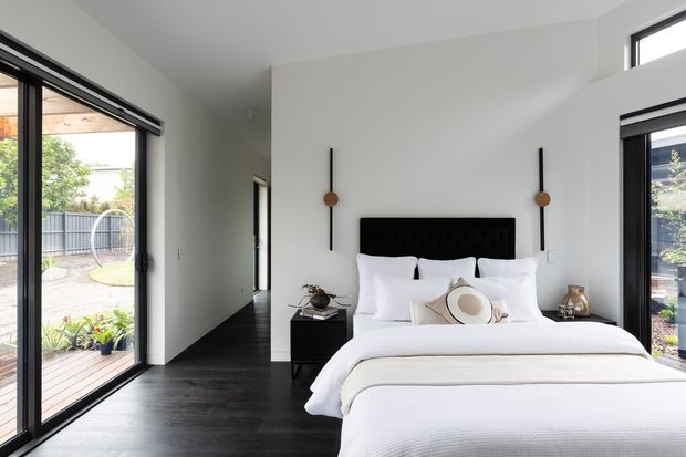Bright contemporary bedroom with a large upholstered black bedhead, crisp white bedding, dark timber flooring, white walls, and floor-to-ceiling glazed doors opening toward an outdoor area. Minimal black-and-brass pendant lights flank the bed, creating a refined, high-contrast interior.