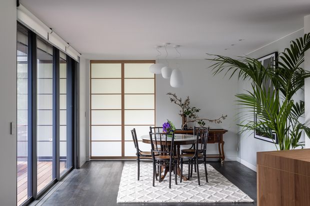 Bright, contemporary dining room with a round wooden dining table and six dark timber chairs arranged on a light patterned rug. Full-height black-framed glazed doors line one side, while a translucent timber-framed sliding screen, sculptural pendant lights, a timber sideboard, framed wall art, and a large indoor palm complete the space. The room has white walls and ceiling with dark timber flooring.