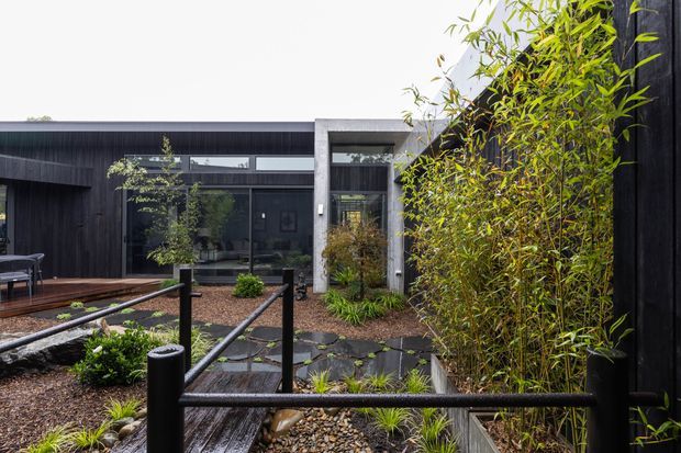 A contemporary dark-clad courtyard viewed from an elevated exterior walkway or deck. The building has charcoal vertical cladding, large dark-framed glazing, and a contrasting concrete-framed entry or courtyard opening. Timber decking, gravel, stepping stones, raised planting beds, ornamental grasses, and dense bamboo create a landscaped, Japanese-influenced outdoor setting. Black metal railings and posts define the walkway.