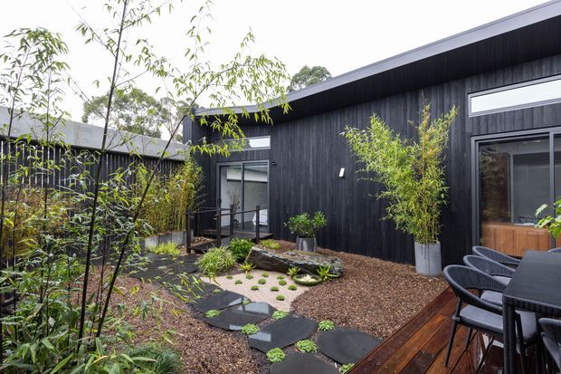 A landscaped courtyard beside a dark timber-clad contemporary building, featuring a timber deck with outdoor dining furniture, stepping stones through gravel, raised planting beds, bamboo and other greenery, and large glazed doors and windows.