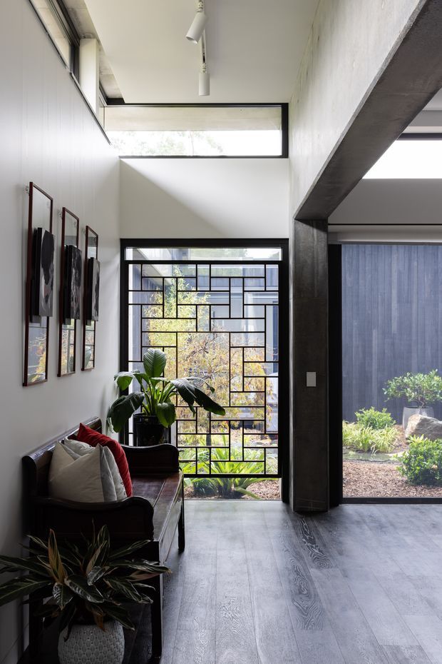 A bright, contemporary residential entry or living space with a tall glazed opening featuring a distinctive black geometric metal grille. The room has pale walls, dark structural framing, grey-toned timber flooring, framed artwork, indoor plants, and a dark leather bench or sofa with cushions. High-level clerestory glazing and minimalist track lighting bring in abundant natural light, while a landscaped courtyard is visible beyond the glazing.