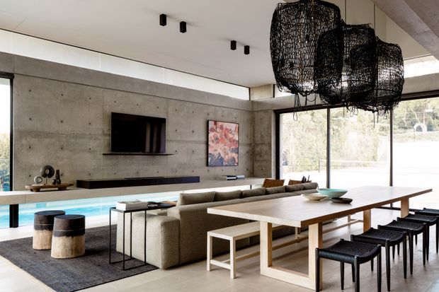 A spacious open-plan contemporary interior combining a living room and dining area. A concrete feature wall with built-in television and linear fireplace faces a large sectional sofa, while a long timber dining table with black stools sits in the foreground. Full-height glazing overlooks a swimming pool and landscaped exterior, and a large sculptural black woven pendant hangs above the dining table.