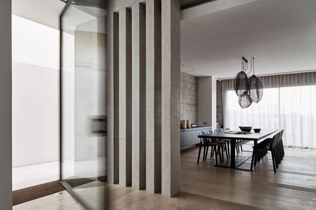 Contemporary open-plan dining area viewed through a glazed entry partition. A dark rectangular dining table with black chairs sits beneath sculptural woven pendant lights, with sheer curtains across large windows. The space features a strong textured concrete vertical screen, exposed concrete wall surfaces, dark built-in cabinetry, and warm timber flooring, creating a refined, neutral-toned interior.
