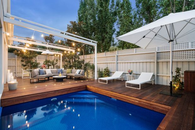 A landscaped residential backyard courtyard centred on a vivid blue swimming pool, surrounded by a timber deck. A pergola with warm festoon lighting shelters an outdoor lounge, while two white sun loungers and a large white umbrella occupy the opposite side. Pale privacy fencing, lattice screening, potted plants and mature trees enclose the space, creating an inviting evening entertaining area.