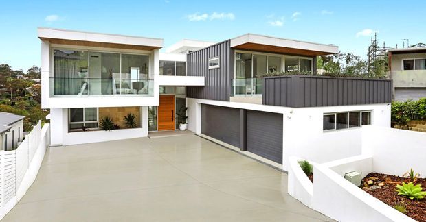 Exterior view of a contemporary multi-level residence with a split modern facade. The home features crisp white rendered walls, dark vertical cladding, broad floor-to-ceiling glazing, projecting flat roof forms, balconies, and an integrated double garage. A large pale concrete driveway and white boundary walls dominate the foreground, with neighbouring suburban homes visible around the property.