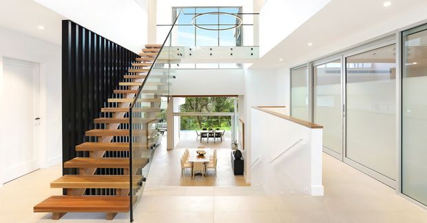 Bright contemporary multi-level interior featuring an open stairway with timber floating treads, a black vertical-slat feature wall, glass balustrades, and a mezzanine overlooking a dining area. White walls, pale flooring, extensive glazing, and a double-height void create an airy, refined space.