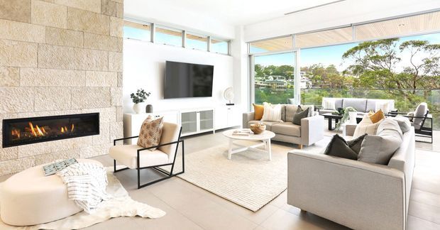 Bright contemporary living room with large floor-to-ceiling windows overlooking trees and neighbouring homes. The room features a full-height pale stone feature wall with a built-in linear fireplace, wall-mounted television, pale built-in media cabinetry, neutral upholstered sofas and armchairs, a light rug, and a white coffee table. An outdoor terrace or balcony is visible beyond the glazing.