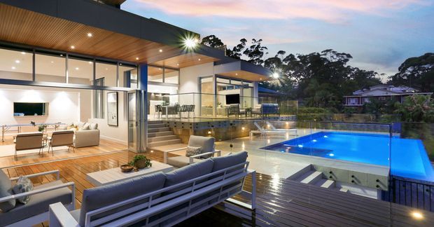 A contemporary multi-level residence photographed at dusk, with extensive floor-to-ceiling glazing opening onto a timber deck and illuminated swimming pool. The outdoor entertaining area includes lounge seating, a dining setting, an outdoor fireplace, and a spa beside the pool. Warm timber soffits and decking contrast with dark metal framing, while the interior living and dining spaces are visible through the glazing. Dense trees and neighbouring homes form the background.