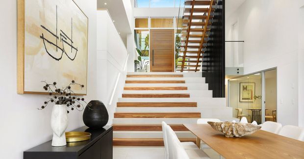 Bright contemporary open-plan interior centred on a sculptural staircase with white risers and warm timber treads. The stair rises beneath a double-height void with a skylight, timber slatted balustrade and black vertical screen. A dining table and chairs occupy the foreground, while a minimalist console with artwork and decorative objects lines the wall.