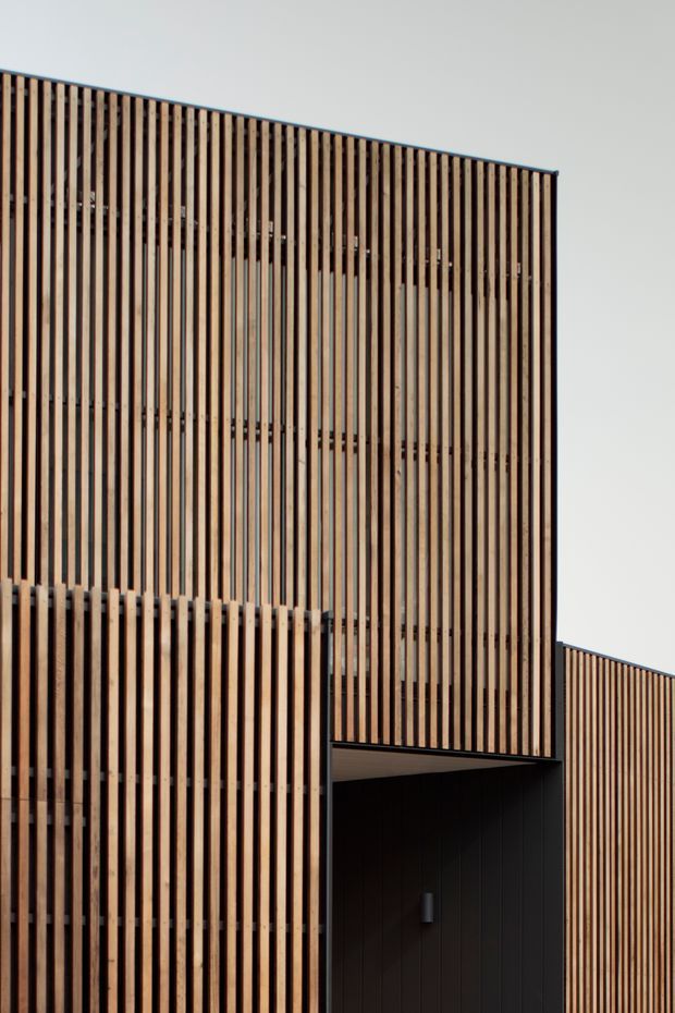 Close-up exterior view of a contemporary multi-level building facade clad in closely spaced vertical timber slats, with dark recessed framing and a deep sheltered entry or opening. The slatted screen creates a strong rhythmic, semi-transparent facade, with a second matching wing visible to the right against a plain sky.