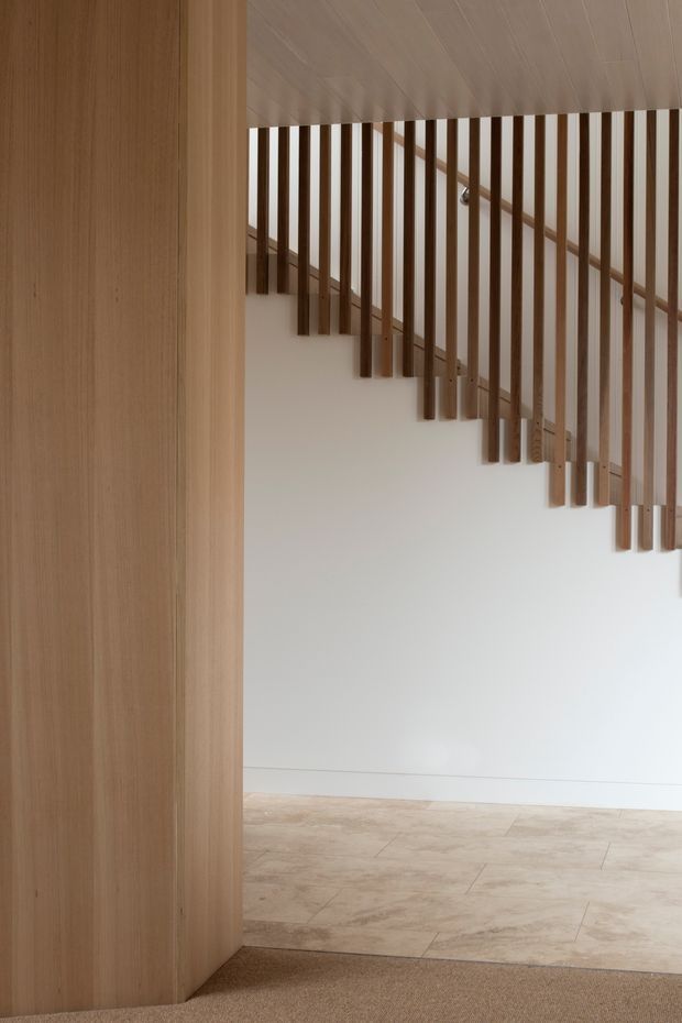 A minimalist interior view of a stairway, framed by a tall light timber-clad wall or joinery panel. The staircase has slim vertical timber balusters following the stair slope, with a pale timber ceiling and handrail, white walls, pale stone or tile flooring, and a neutral carpeted foreground. The composition emphasizes clean lines, natural materials, and filtered light.