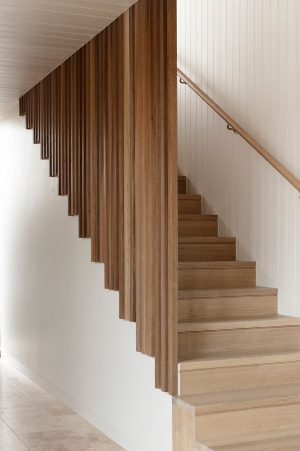 Interior architectural photograph of a minimalist stairway. A warm light-wood staircase rises beside a white vertically panelled wall, with a simple timber handrail and striking vertical timber slat screen forming the balustrade or stair enclosure. The adjacent floor appears to be pale stone or tile, and the composition is bright, clean, and highly architectural.