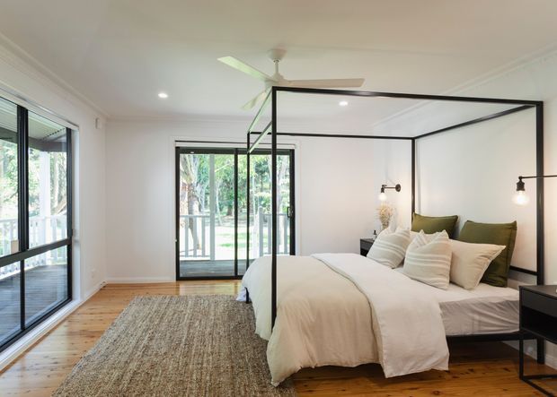 Bright, spacious bedroom with a black metal four-poster canopy bed dressed in white linen bedding and olive-green accent cushions. The room has warm timber flooring, a large neutral textured rug, white walls with decorative cornices, expansive black-framed glazing and doors opening to a leafy outdoor setting, recessed ceiling lights, wall-mounted bedside sconces, and a ceiling fan.
