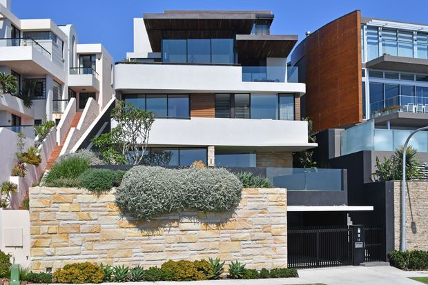 Street-facing exterior view of a contemporary multi-storey residence in a dense coastal-style urban neighbourhood. The house has crisp white rendered volumes, extensive dark-framed glazing, projecting balconies and terraces, dark timber soffits, a rust-toned metal-clad upper wall, and a low sandstone retaining wall with landscaped planting in front. A black garage gate and driveway are visible at ground level.