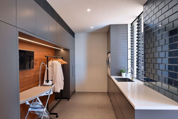 A sleek contemporary dressing and laundry-style utility room with extensive dark grey built-in cabinetry. The room includes a timber-lined open dressing niche with hanging clothes, a fold-out ironing board, a long white countertop with an inset sink, and a dramatic dark blue-grey tiled feature wall. Narrow louvred windows provide filtered daylight along the right side.