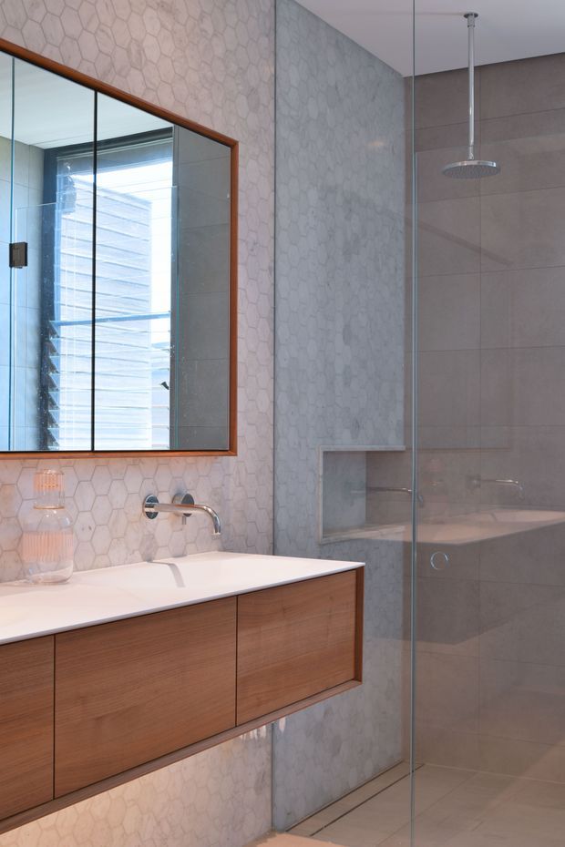 A refined contemporary bathroom featuring a floating timber vanity with a white benchtop, wall-mounted chrome tapware, a large timber-framed mirror, pale hexagonal wall tiles, and a frameless glass shower enclosure with large-format grey tiles and a recessed niche. The space is softly lit and uses a restrained neutral palette with warm timber accents.
