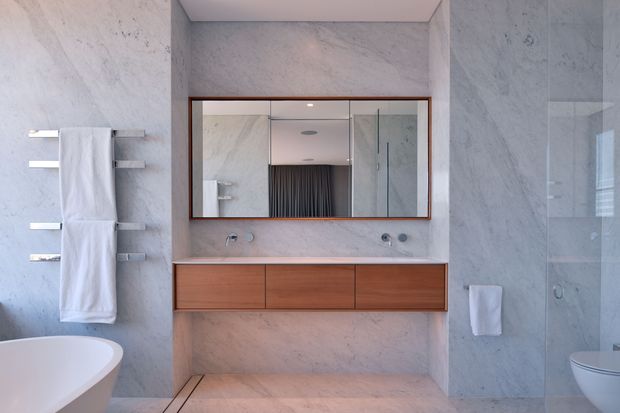 Contemporary bathroom featuring full-height pale marble-look wall and floor surfaces, a floating warm timber vanity with integrated twin basins and wall-mounted fixtures, a large framed mirrored cabinet, chrome heated towel rail, freestanding white bathtub, and a glazed shower enclosure. The space is clean, refined, and softly lit.