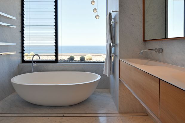 A refined contemporary bathroom with a freestanding white oval bathtub positioned beside a large window overlooking the ocean. The room features grey stone-look wall and floor finishes, a timber floating vanity with an integrated pale benchtop, wall-mounted chrome tapware, a frameless mirror, and a vertical towel rail. Horizontal louvres provide privacy and filtered daylight, while pendant globe lights add a subtle decorative element.