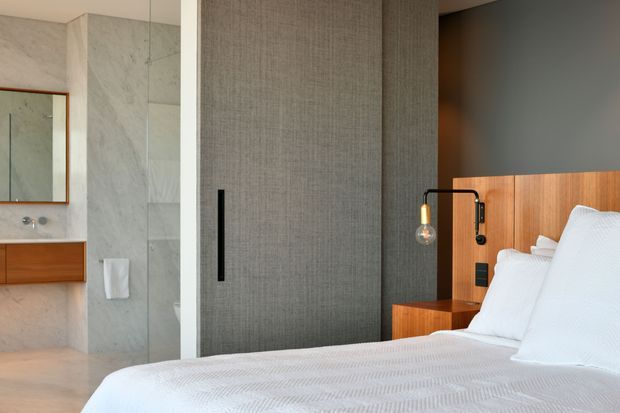 Contemporary bedroom with a partially visible ensuite bathroom. The room features a white textured bed, warm timber bedhead and bedside joinery, a dark grey wall, a full-height textured grey wardrobe or sliding door, and a pendant bedside light. The ensuite includes pale stone-look wall surfaces, a timber vanity, mirror, and glass shower enclosure.