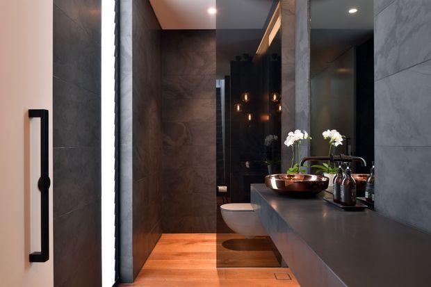 A dark, luxurious contemporary bathroom viewed from the entry. Large grey stone-look wall tiles surround a walk-in shower, with warm timber flooring, a wall-hung toilet, and a long dark floating vanity with an integrated basin and brushed metal tapware. A large mirror, pendant reflections, orchids, and black accessories add refinement and contrast.