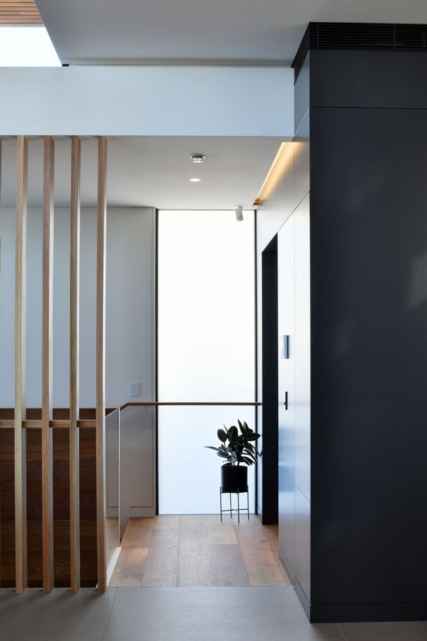 A modern residential hallway or landing with pale timber flooring, white walls and ceiling, a black full-height built-in joinery wall, and a vertical timber batten screen partially enclosing a stairwell. Minimal black-and-white palette is softened by warm timber accents and a potted plant. A bright glazed or open area is visible at the end of the corridor, with recessed and linear lighting integrated into the ceiling and cabinetry.