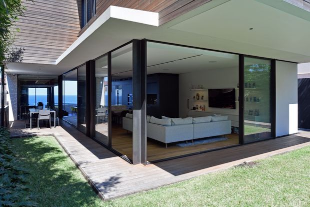 Exterior view of a contemporary multi-level residence with extensive black-framed floor-to-ceiling glazing opening onto a timber deck and lawn. The covered outdoor area connects to a bright living room with a large sectional sofa, television, and built-in shelving. A dining area and expansive ocean view are visible beyond the glazed corridor. Timber cladding and soffit lining combine with crisp white walls and dark metal frames.